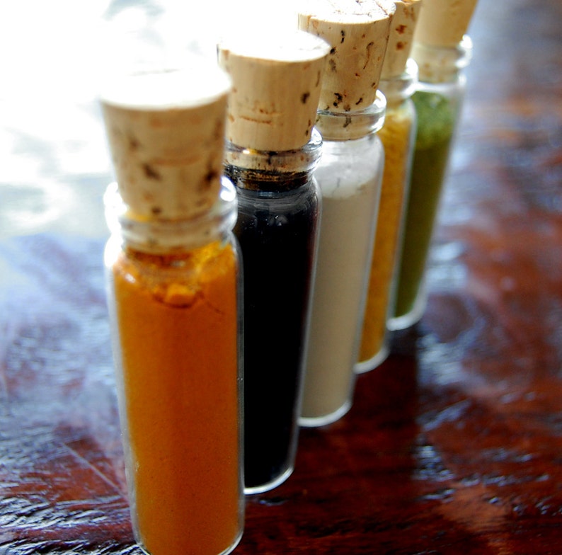 May include: Five small glass bottles with cork tops, filled with different colored powders. The bottles are lined up in a row on a wooden surface. The powders are yellow, black, white, light brown, and green.