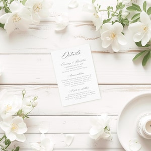 May include: A white card with black text detailing the ceremony and reception for a wedding. The card includes the time, location, and a note about children. The card is on a white wooden surface with white flowers.