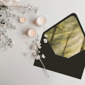 May include: A black envelope with a forest scene printed on the inside flap. The envelope is surrounded by white flowers and candles on a white surface.