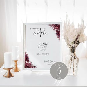 Peut inclure: Une pancarte blanche avec le texte "The Perfect Match" et un dessin d'une boîte d'allumettes avec des cœurs. La pancarte comprend également les noms "Erica & Stephen" et la date "23.11.22". La pancarte est décorée d'illustrations florales bordeaux. Un cercle avec le texte "Includes 5 Sizes" se trouve dans le coin inférieur droit.