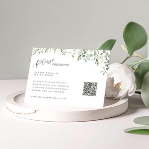 以下が含まれることがあります： 緑の葉と白い花が描かれた白いRSVPカード。テキストには「ご返信をお願いします」と「23.10.2XXXまでにご返信ください」と記載されています。カードにはQRコードとウェブサイトアドレス「WWW.OLIVIABMASON.COM」も記載されています。