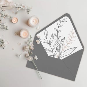 May include: A gray envelope with a white liner featuring a black and brown line drawing of leaves and branches. The envelope is lying on a white surface with white flowers and candles.