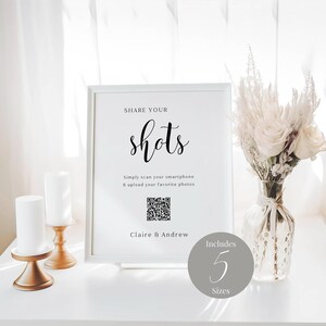 May include: White sign with black text that reads "SHARE YOUR shots Simply scan your smartphone & upload your favorite photos Claire & Andrew Includes 5 Sizes". A QR code is included on the sign. The sign is sitting on a white surface with a vase of white flowers and two candlesticks.