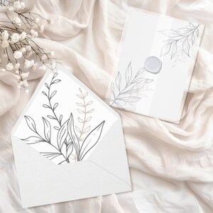 May include: A white envelope with a floral design on the front. The envelope is lying on a white fabric surface. The floral design is in shades of gray and brown.