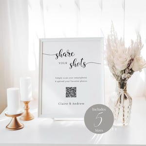 Pode incluir: Um sinal branco com texto preto que diz "share your shots". O sinal também inclui um código QR e o texto "Simply scan your smartphone & upload your favorite photos". O sinal está sobre uma mesa branca com um vaso de flores brancas e duas velas.