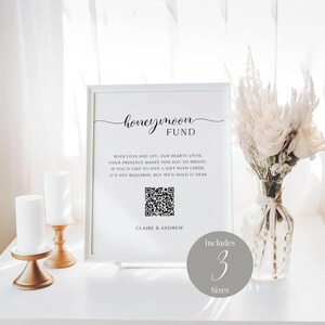 May include: A white sign with black text that reads "honeymoon fund" with a QR code and the text "with love and joy, our hearts unite. your presence makes this day so bright. if you'd like to give a gift with cheer, it's not required, but we'd hold it dear. claire & andrew". The sign is on a white table with a vase of white flowers and two gold candle holders.