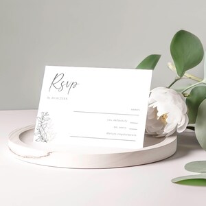 以下が含まれることがあります： 白いRSVPカードに手書きの「RSVP」と花のイラストが描かれています。カードには、ゲストが参加の可否と食事に関する要望を記入する欄があります。「by 22.03.XXXX」というテキストがRSVPの下に印刷されています。