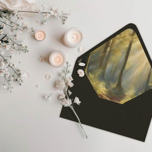 May include: A black envelope with a forest scene painted on the front. The scene depicts a sunlit path through a forest with tall trees and a hazy atmosphere. The envelope is surrounded by white flowers and candles.