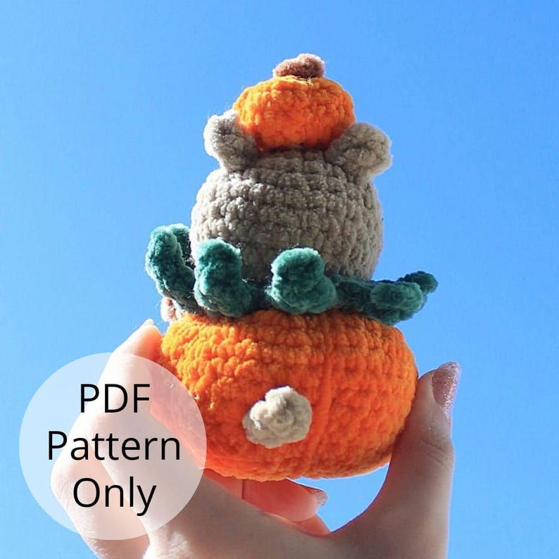 May include: A crocheted pumpkin stack with a gray, fuzzy pumpkin on top, a green leafy layer in the middle, and an orange pumpkin on the bottom.