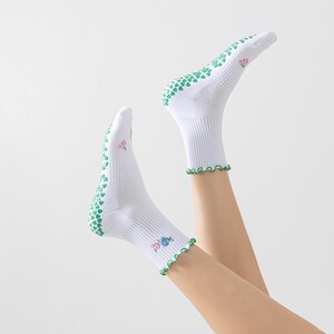 Puede incluir: Calcetines blancos hasta el tobillo con agarres en forma de coraz&oacute;n verde en la parte inferior y un ribete festoneado verde. Los calcetines tienen un dise&ntilde;o floral en el lateral.