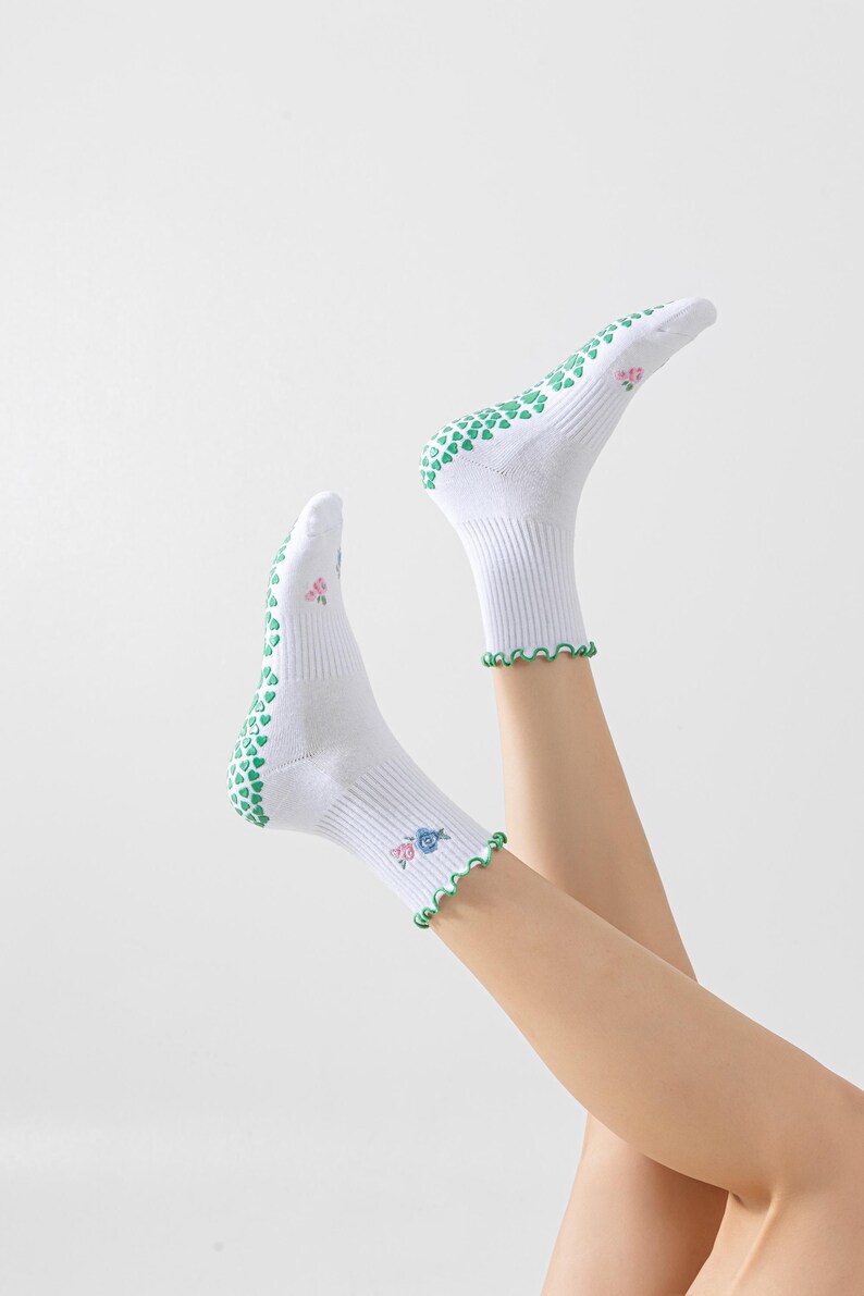 Puede incluir: Calcetines blancos hasta el tobillo con agarres en forma de coraz&oacute;n verde en la parte inferior y un ribete festoneado verde. Los calcetines tienen un dise&ntilde;o floral en el lateral.