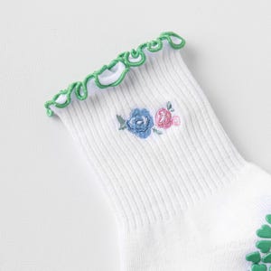 Puede incluir: Calcetines blancos con un borde superior de volantes verdes y un peque&ntilde;o dise&ntilde;o floral bordado en azul y rosa. Los calcetines tienen agarres antideslizantes verdes en la parte inferior.