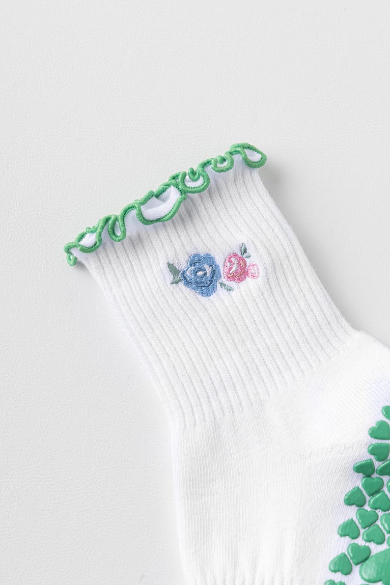 Puede incluir: Calcetines blancos con un borde superior de volantes verdes y un peque&ntilde;o dise&ntilde;o floral bordado en azul y rosa. Los calcetines tienen agarres antideslizantes verdes en la parte inferior.