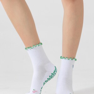 Puede incluir: Calcetines blancos hasta el tobillo con agarres antideslizantes verdes en la parte inferior y un ribete verde con volantes alrededor de la parte superior. Los calcetines tienen un peque&ntilde;o dise&ntilde;o floral en la parte delantera.
