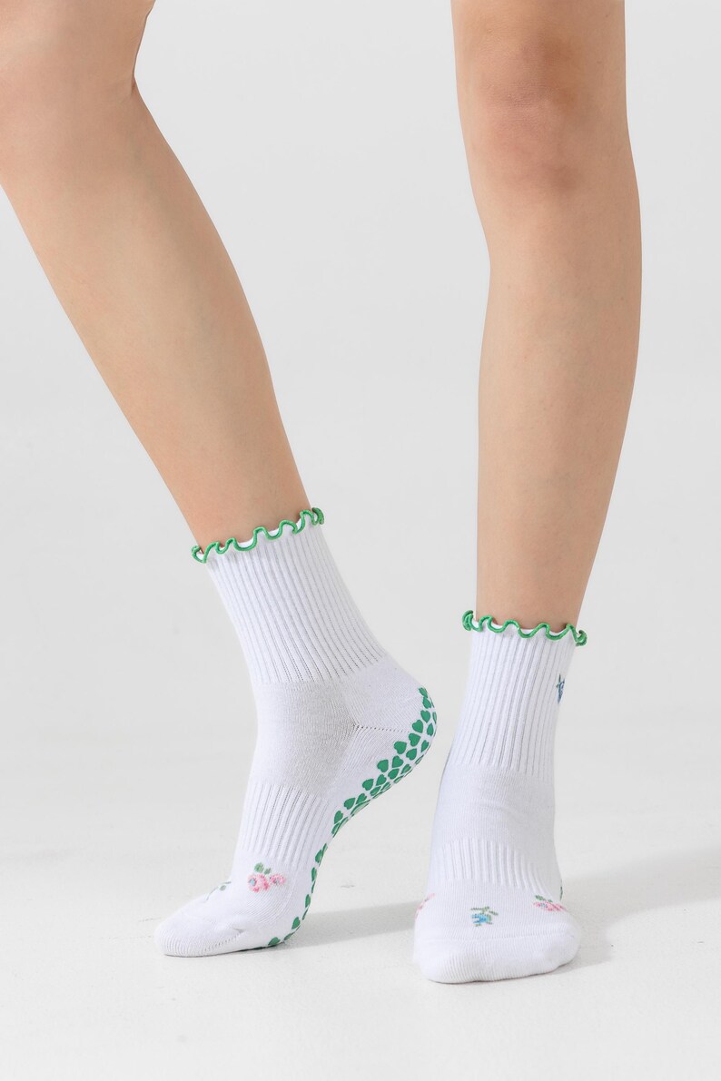 Puede incluir: Calcetines blancos hasta el tobillo con agarres antideslizantes verdes en la parte inferior y un ribete verde con volantes alrededor de la parte superior. Los calcetines tienen un peque&ntilde;o dise&ntilde;o floral en la parte delantera.
