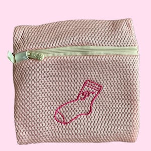 Puede incluir: Una bolsa de lavandería de malla rosa con cierre de cremallera y una media rosa bordada en la parte delantera.