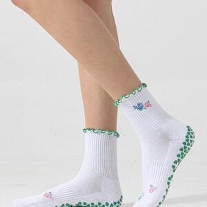Puede incluir: Calcetines blancos hasta el tobillo con puntos antideslizantes verdes en la parte inferior y un volante verde y blanco en la parte superior. Los calcetines tienen un peque&ntilde;o dise&ntilde;o floral bordado en el lateral.