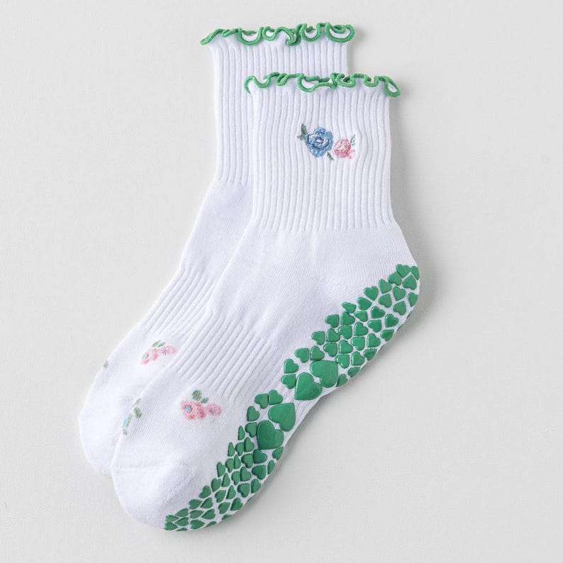 Puede incluir: Calcetines blancos hasta el tobillo con puntos antideslizantes verdes en la parte inferior y un dise&ntilde;o de bordado floral. Los calcetines tienen un ribete verde con volantes en la parte superior.