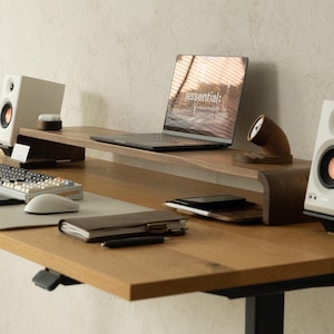 May include: A wooden desk with a laptop displaying "essential;" and a keyboard. The desk has a monitor stand, speakers, a notebook, and a pen. The desk is made of light-colored wood.