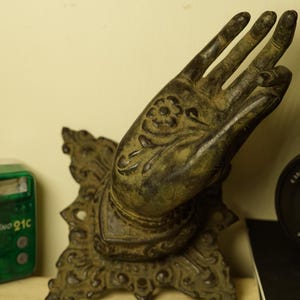 Puede incluir: Escultura de mano de color bronce con intrincados detalles florales en la palma. La mano está montada sobre una base decorativa con patrones ornamentados. La escultura tiene una apariencia desgastada, lo que sugiere una pieza antigua o vintage.