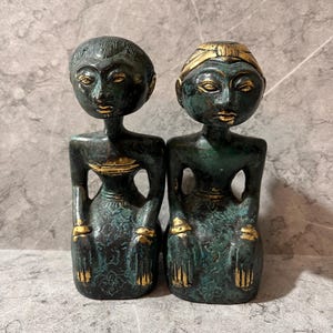 Könnte beinhalten: Zwei bronzefarbene Statuen sitzender Figuren mit stilisierten Merkmalen. Die Figuren haben eine dunkelgrüne Patina mit goldenen Akzenten auf Kopfbedeckung, Kleidung und Händen. Die Statuen werden nebeneinander ausgestellt.
