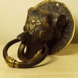 Peut inclure: Un heurtoir de porte en forme de tête d'éléphant en laiton orné, avec une poignée en anneau. La tête de l'éléphant est ornée de gravures complexes et d'une plaque arrière circulaire. Le heurtoir a une patine sombre.