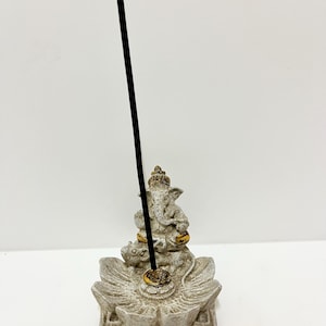 May include: A grey and gold incense holder depicting the Hindu deity Ganesha. The holder is shaped like a lotus flower and holds a black incense stick. The sculpture has gold detailing. The incense holder is made of stone.