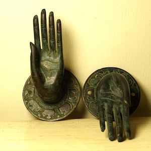 Puede incluir: Dos apliques de pared con forma de mano en tono bronce. Cada mano está montada sobre una base circular con un diseño ornamental, similar a una flor. Las manos están en un gesto de bienvenida, con los dedos extendidos.