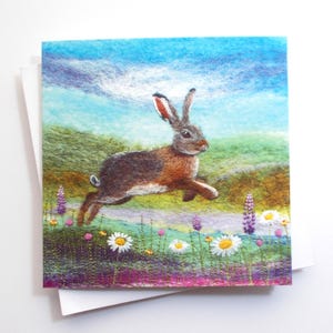 May include: A card with a felted image of a hare leaping across a meadow. The hare is brown with orange and white markings, set against a backdrop of green hills and a blue sky. White daisies and purple flowers are in the foreground.