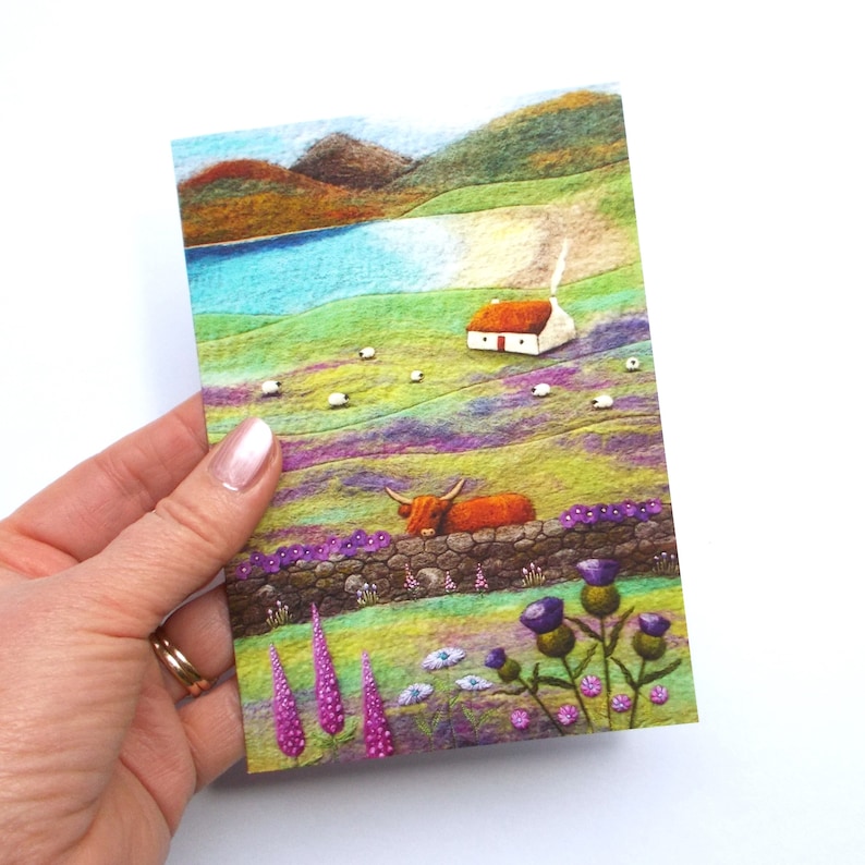 A vibrant landscape greetings card depicting a Highland cow peeking over on a drystone wall. The scene includes a small white cottage, sheep grazing in a field, and a cove with mountains in the background. Purple and pink flowers are in the foreground. The original picture was created using wool felt and embroidery and has been reproduced onto quality card. Card is blank and comes with a plain white envelope.