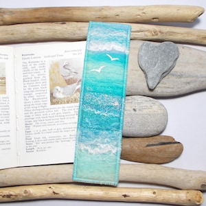 May include: A handmade bookmark featuring a seascape design. The bookmark has a turquoise and white color scheme, depicting ocean waves and two white seagulls. The bookmark is 16 inches (41 cm) long.