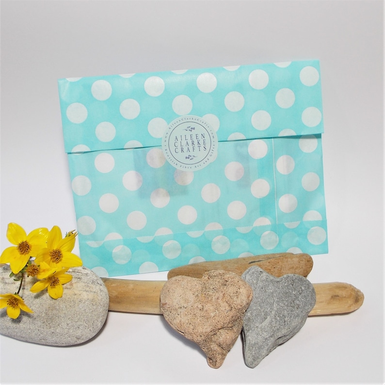 May include: A turquoise polka dot gift bag with a clear window and a round sticker that reads "Aileen Clarke Crafts". The bag is next to a gray stone, two heart-shaped stones, driftwood, and yellow flowers.
