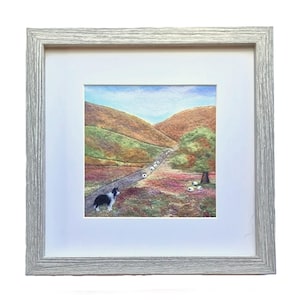 May include: A framed needle felted artwork depicting a black and white border collie herding a flock of white sheep up a winding path in a green and brown hilly landscape.