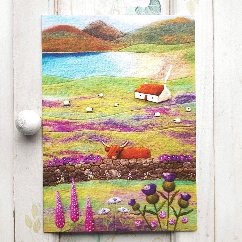 A vibrant landscape greetings card depicting a Highland cow peeking over on a drystone wall. The scene includes a small white cottage, sheep grazing in a field, and a cove with mountains in the background. Purple and pink flowers are in the foreground. The original picture was created using wool felt and embroidery and has been reproduced onto quality card. Card is blank and comes with a plain white envelope.