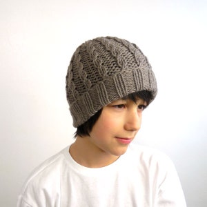 May include: A grey knitted beanie with a cable knit pattern. The beanie is being worn by a young person with dark hair.
