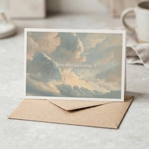 Può includere: Un biglietto d'auguri rettangolare con l'immagine stampata di un cielo nuvoloso nei toni tenui del blu, bianco e pesca. Il testo "blue skies are coming" è centrato sul biglietto in un carattere bianco senza grazie. Sotto è posizionata una busta di carta marrone.