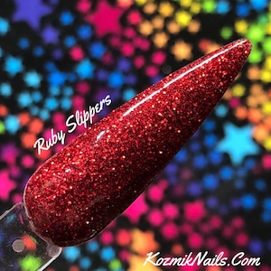 May include: A close-up of a red glitter nail polish with the text "Ruby Slippers" on the side. The polish is on a clear nail tip.