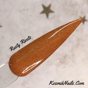 May include: A close-up of a nail polish color called "Rusty Rivets" with a golden shimmer finish. The polish is on a clear nail tip.