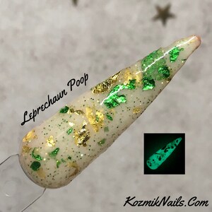 May include: A white nail polish with green and gold glitter and flakes. The polish glows green in the dark. The text "Leprechaun Poop" is written on the nail.