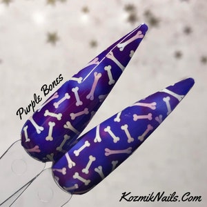 May include: Two long, pointed, purple nail tips with a white bone pattern. The nail tips are decorated with a white bone pattern on a purple background. The text "Purple Bones" is written on the left side of the image.