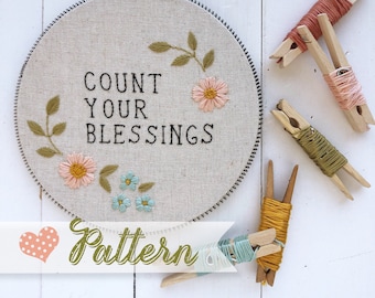 May include: Embroidery hoop with the phrase "Count Your Blessings" stitched in black thread. The hoop is surrounded by colorful embroidery floss on wooden clothespins. The phrase "Pattern" is written in green on a white heart-shaped background.