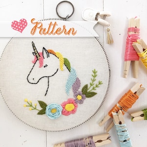 May include: Embroidered hoop art featuring a white unicorn with a rainbow mane and tail, surrounded by colorful flowers and green leaves. The hoop is labeled "Pattern" with a heart.