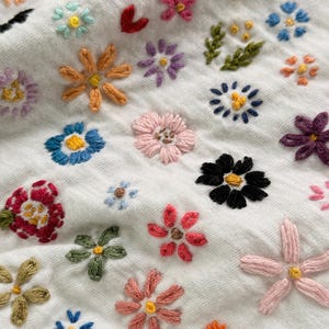 May include: Close-up of white fabric decorated with various embroidered flowers. The flowers are in shades of orange, pink, blue, and purple, with yellow centres. The embroidery gives a textured, handmade look.