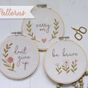 Peut inclure: Trois cerceaux à broder avec des citations inspirantes et des motifs floraux. Les cerceaux sont en bois et ont un fond en tissu blanc. Les citations sont "carry on", "don't give up", et "be brave".