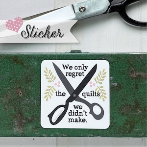 May include: Square sticker with the text "We only regret the quilts we didn't make" and a pair of black scissors. The sticker has a white background with floral accents and is on a green surface. A banner with the word "Sticker" is in the upper left.