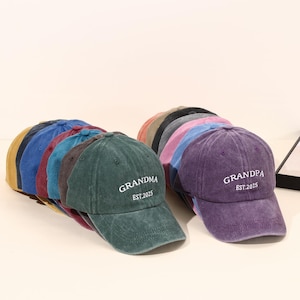 May include: A selection of baseball caps in shades like green, purple, and blue. Each cap is embroidered with either "GRANDMA" or "GRANDPA" and "EST.2025". The caps are displayed in a row, showcasing a range of colours.