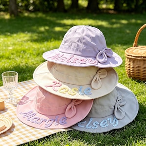 Può includere: Una pila di quattro cappelli da sole in lavanda, beige, rosa e grigio chiaro. Ogni cappello presenta un fiocco laterale e nomi ricamati sulla tesa: Nelsey, Rosie, Margie ed Elsey. I cappelli sono realizzati in tessuto con tesa larga.