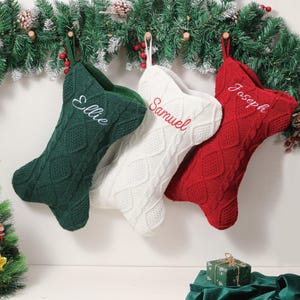 Op de afbeelding: Drie kerstsokken in groen, wit en rood hangen aan een slinger. Elke sok heeft een kabelgebreid ontwerp en een naam geborduurd in een contrasterende kleur: "Ellie", "Samuel" en "Joseph".