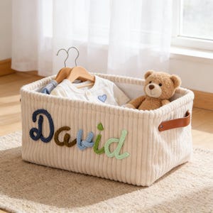 Może przedstawiać: Beżowy kosz do przechowywania z sztruksu z napisem "David" w niebieskich, brązowych i zielonych literach. Kosz zawiera ubranka dla dzieci na drewnianych wieszakach i małego misia. Posiada brązowe skórzane uchwyty.