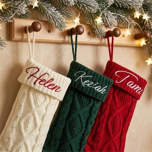 Può includere: Tre calze natalizie in maglia appese a un supporto in legno. Le calze sono color crema, verde scuro e rosso, ognuna con un motivo a trecce e un risvolto. I nomi Helen, Keziah e Tami sono ricamati in corsivo.